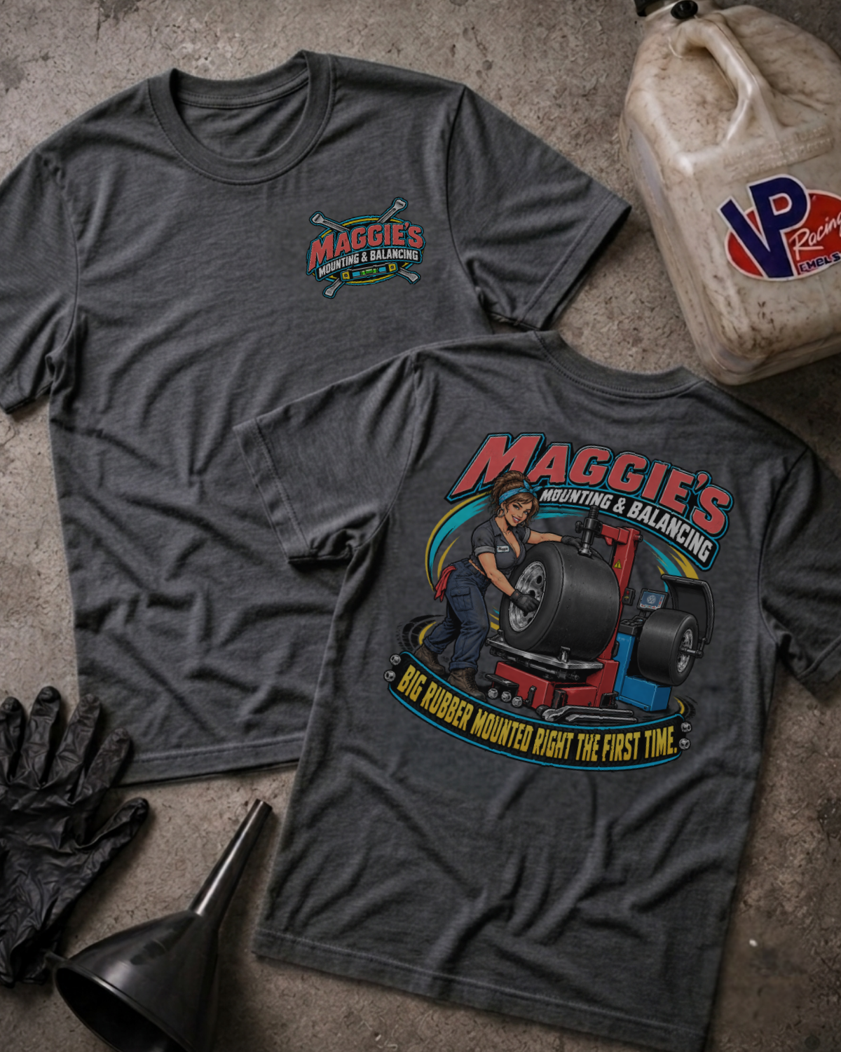 Two dark heather gray graphic T-shirts laid flat on a concrete surface, with a jugs container, gloves, and funnel as styled props. The front shirt shows a small 'Maggie's Mounting & Balancing' logo on the left chest, and the back shirt shows a large colorful graphic of a mechanic working under a tire with the text 'Maggie's Tire Mounting & Balancing' and 'Big Rubber Mounted Right The First Time.