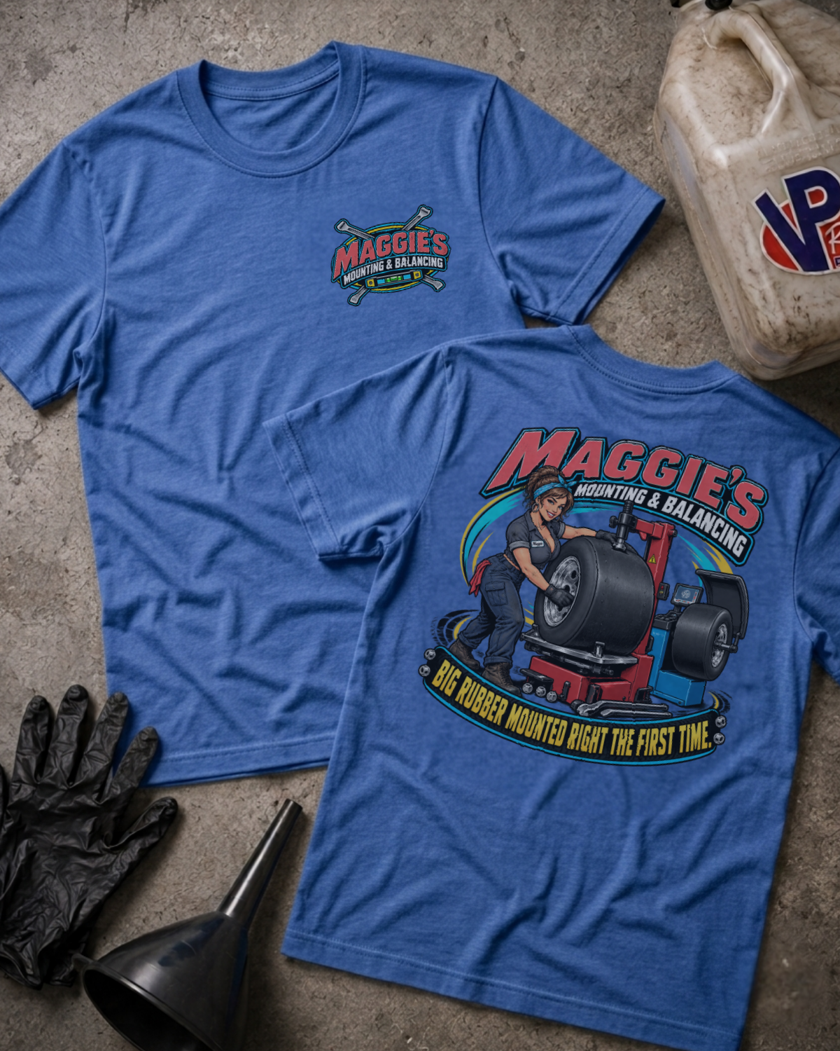 Two blue short-sleeve graphic T-shirts laid flat on a textured concrete surface, with a black oil jug, black gloves, and a black funnel nearby. The front shirt shows a small 'Maggie's Mounting & Balancing' logo on the left chest, and the back shirt shows a large colorful graphic of a mechanic working under a tire on a balancing machine with the text 'Maggie's Mounting & Balancing' and 'Big Rubber Mounted Right the First Time.