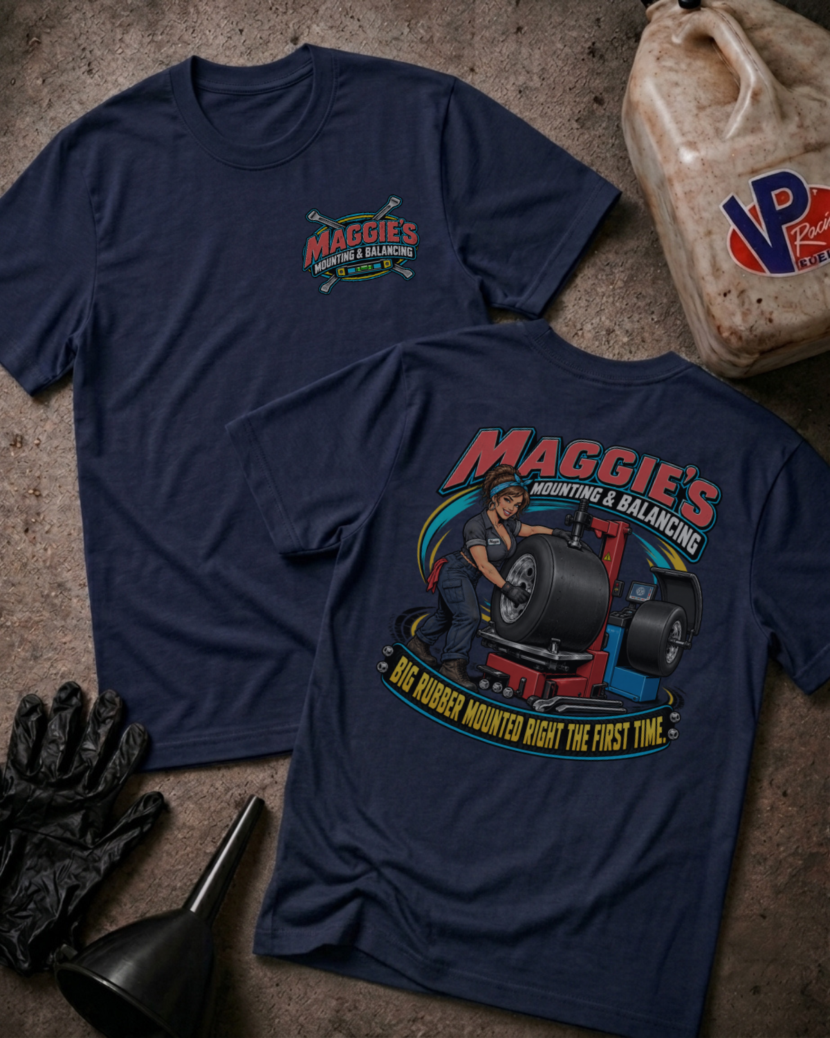 Two navy blue t-shirts displayed on a rustic concrete floor, one showing the small front chest logo and the other showing the large back graphic for 'Maggie's Mounting & Balancing'. The shirts are styled with black gloves, a funnel, and a plastic jug of oil.