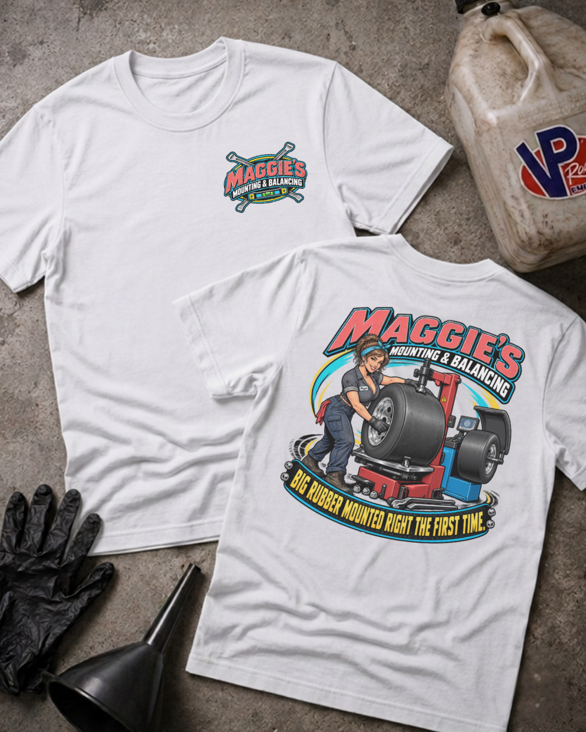 Two white graphic T-shirts laid flat on a concrete surface, showing the front and back designs. The front has a small colorful chest graphic reading 'Maggie's Mounting & Balancing,' and the back has a large racing-themed graphic with a mechanic, tire, and text.
