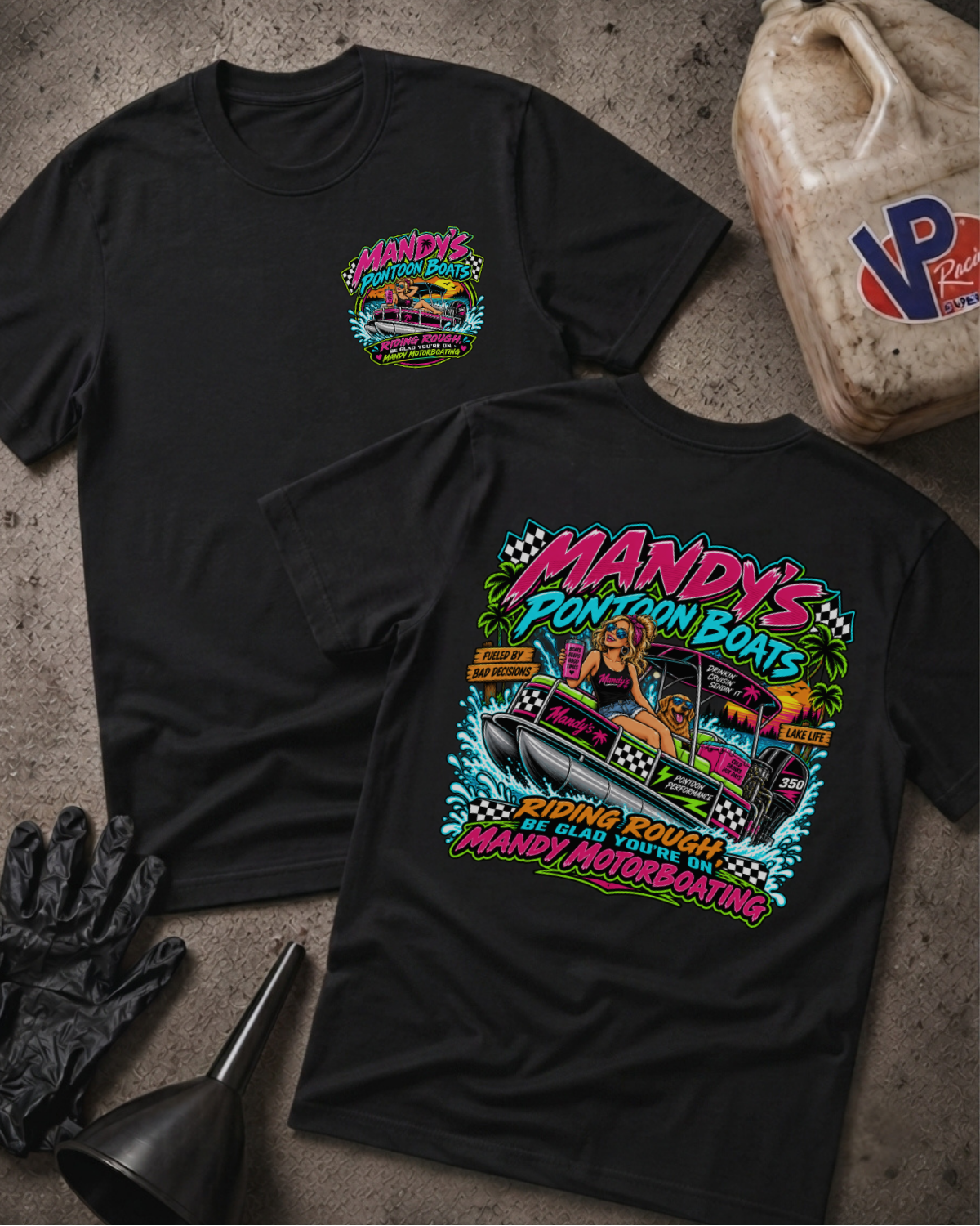 Two black graphic T-shirts laid flat on a textured gray surface, with a large colorful 'Mandys Pontoon Boats' boat illustration on the back and a smaller matching chest graphic on the front. Black gloves and a fuel jug are placed around them as styling props.