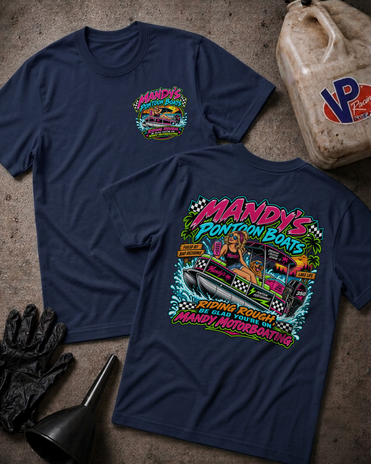 Two navy blue t-shirts displayed on a textured concrete surface with racing-themed props. The front shirt shows a small colorful chest graphic, while the back shirt displays a large, vibrant graphic of a woman in a boat with the text 'MANDY'S PONTOON BOATS' and 'RIDING ROUGH BE GLAD YOU'RE ON MANDY MOTORBOATING'.