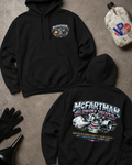 Two black hoodies laid flat on a concrete-style surface, one showing the front chest graphic 'McFARTMAN RACING' logo and the other the large back graphic. A white jug with a blue VP logo and black gloves are placed around it as styled props.