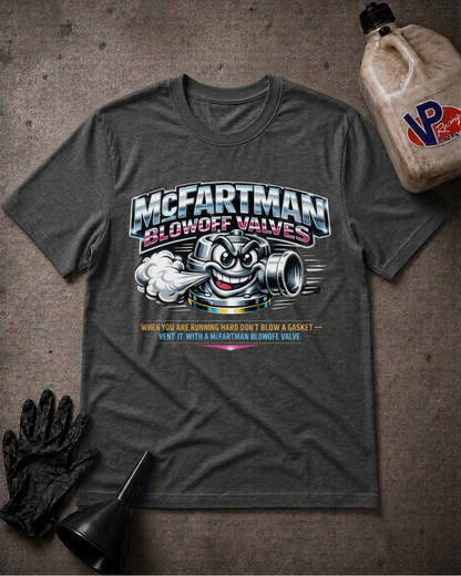 A dark heather gray graphic t-shirt featuring a cartoon exhaust valve and the text 'McFartman Blowoff Valves' with a humorous slogan below, styled flat-lay on a textured gray surface with a black glove, funnel, and a VP Fast & Furious branded bottle.