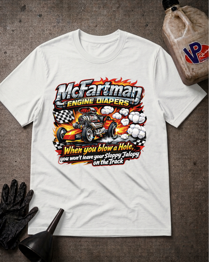 A white graphic t-shirt laid flat on a dark textured surface, surrounded by racing-themed props including a black glove, funnel, and a fuel jug. The shirt features a large graphic with the text 'McFartman Engine Diapers' and a hot rod race car illustration.