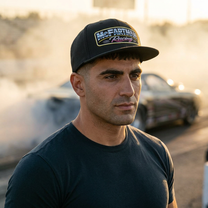 Man wearing a black McFartman Racing snapback hat near the burnout box at a drag strip, tire smoke drifting behind with a drag car in motion, golden hour lighting and gritty race day atmosphere