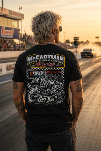 Rear view of man wearing a black McFartman Racing “Get Bent!” drag racing t-shirt at a drag strip during golden hour with burnout smoke, staging lanes, and gritty motorsports atmosphere.