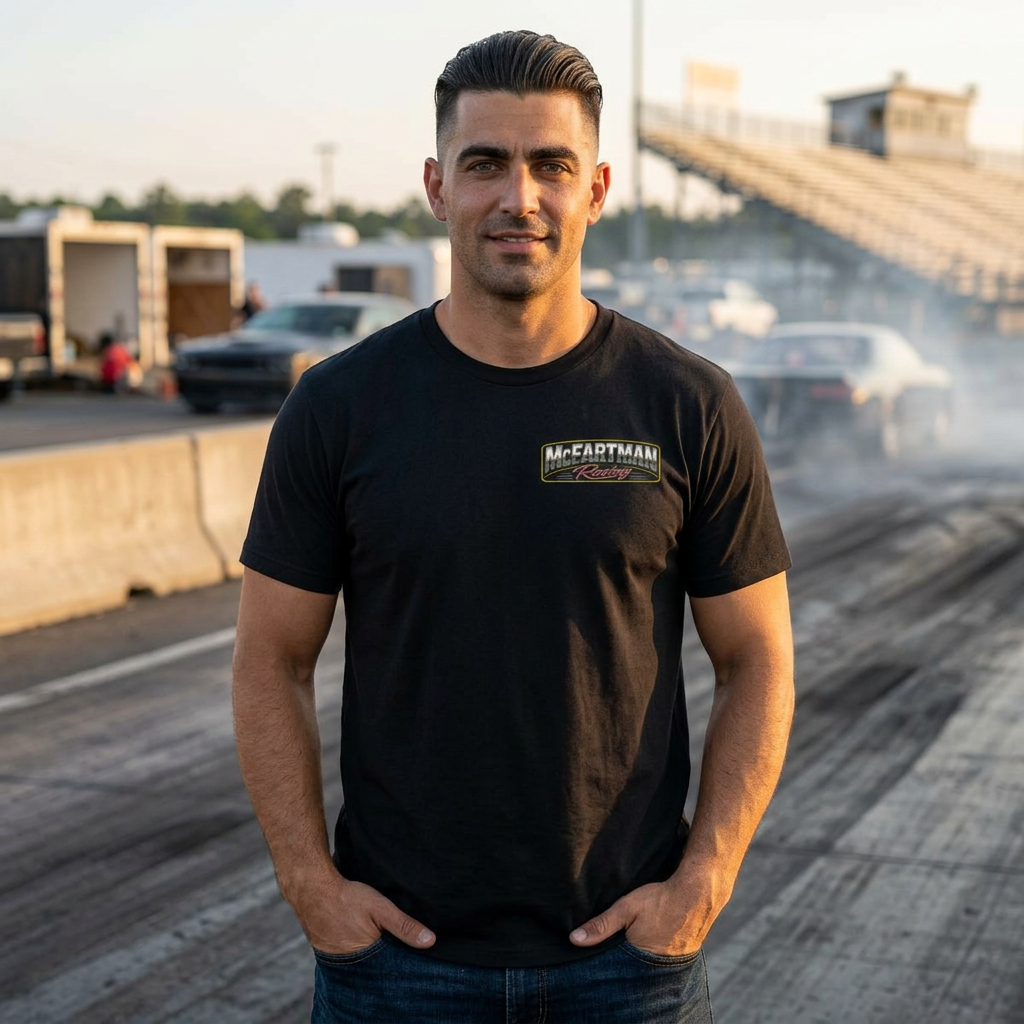 Man wearing a black McFartman Racing drag racing t-shirt at the race track during golden hour with drag cars, burnout haze, and grandstands in the background.