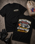 Two black short-sleeve graphic T-shirts laid flat on a textured gray surface, with racing and burrito-themed printed graphics. A large back print reads "McFartman Racing" and "Powered by Nitromethane Burritos," while the front chest has a smaller matching logo.