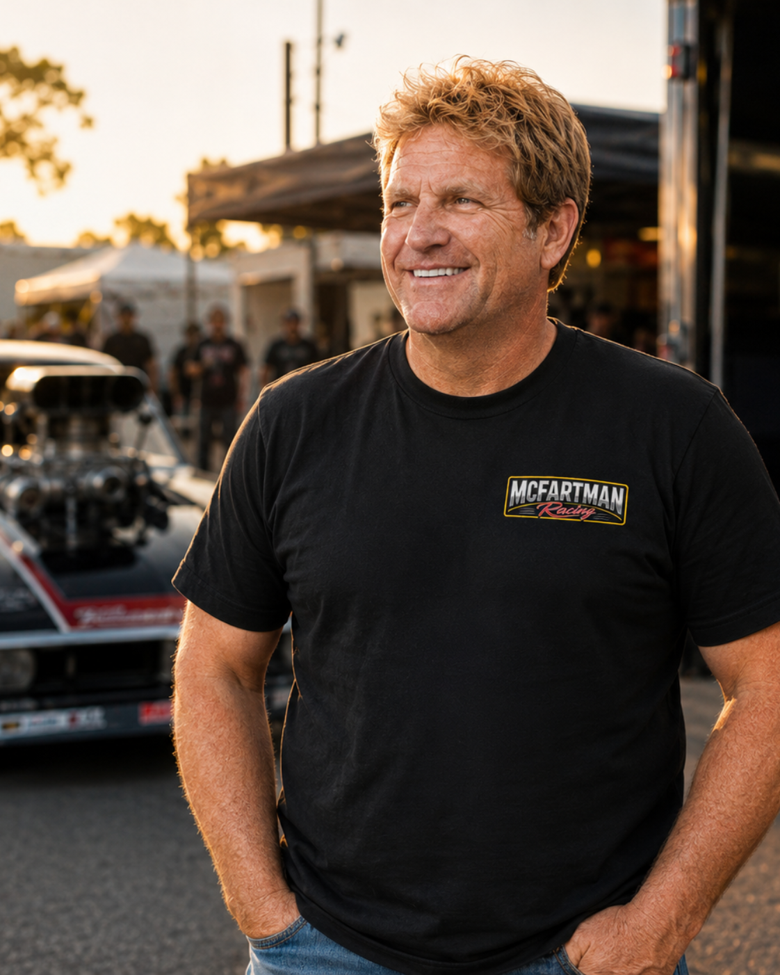 Man wearing a black t-shirt with a McFartman Racing logo standing in front of a race car and trailer.