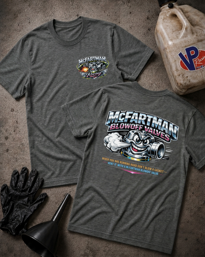 Two heather dark gray graphic T-shirts laid flat on a textured concrete surface, with black gloves and a black funnel in the foreground and a dirty plastic jug in the upper right. The front shirt shows a small chest graphic reading McFartman, and the back shirt shows a large graphic with the same brand name, a cartoon valve mascot, and text about blowing off valves.