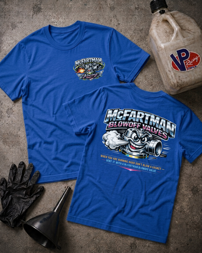 Two blue short-sleeve T-shirts laid flat on a textured gray surface, one showing a small chest graphic and the other showing a large back graphic with the text 'McFartman Blowoff Valves' and a cartoon blower. A plastic jug, black gloves, and a funnel are arranged around them as styling props.