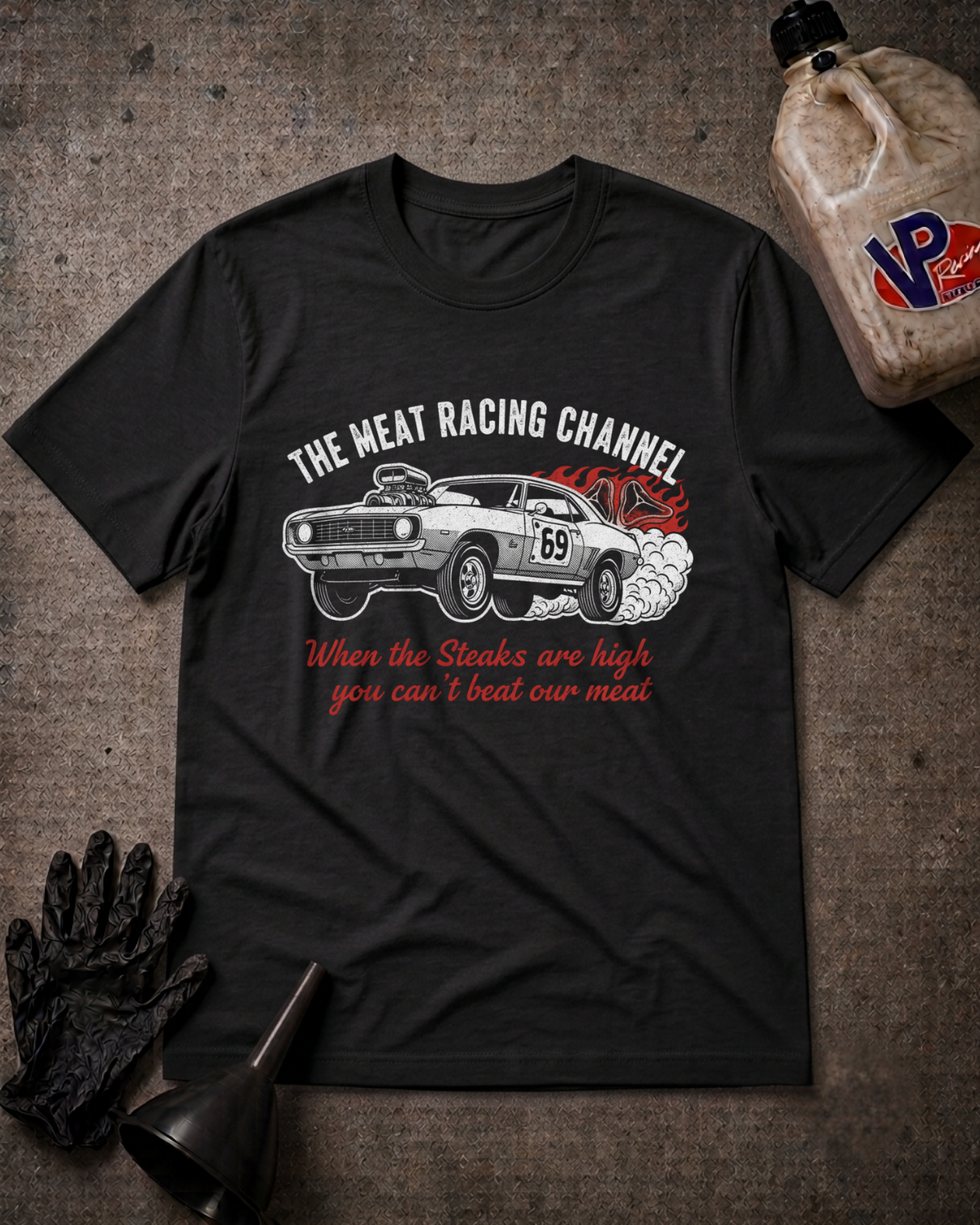 A black graphic t-shirt laid flat on a textured dark background, featuring a white muscle car with red smoke effects and the text 'THE MEAT RACING CHANNEL' and 'When the Steaks are high you can't beat our meat'. A bottle, gloves, and funnel are styled around it as props.