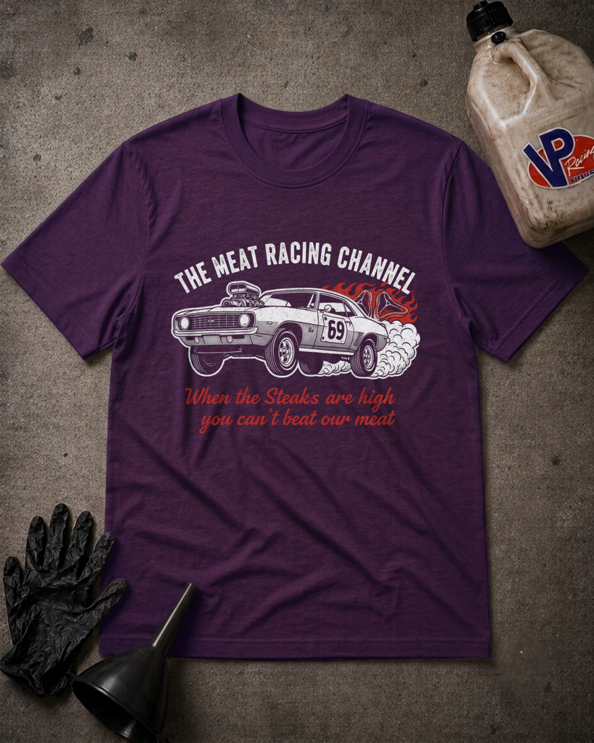 A purple graphic t-shirt featuring a white muscle car with red and white text, laid flat on a gray concrete-textured background with racing-themed props including a bottle, gloves, and funnel.