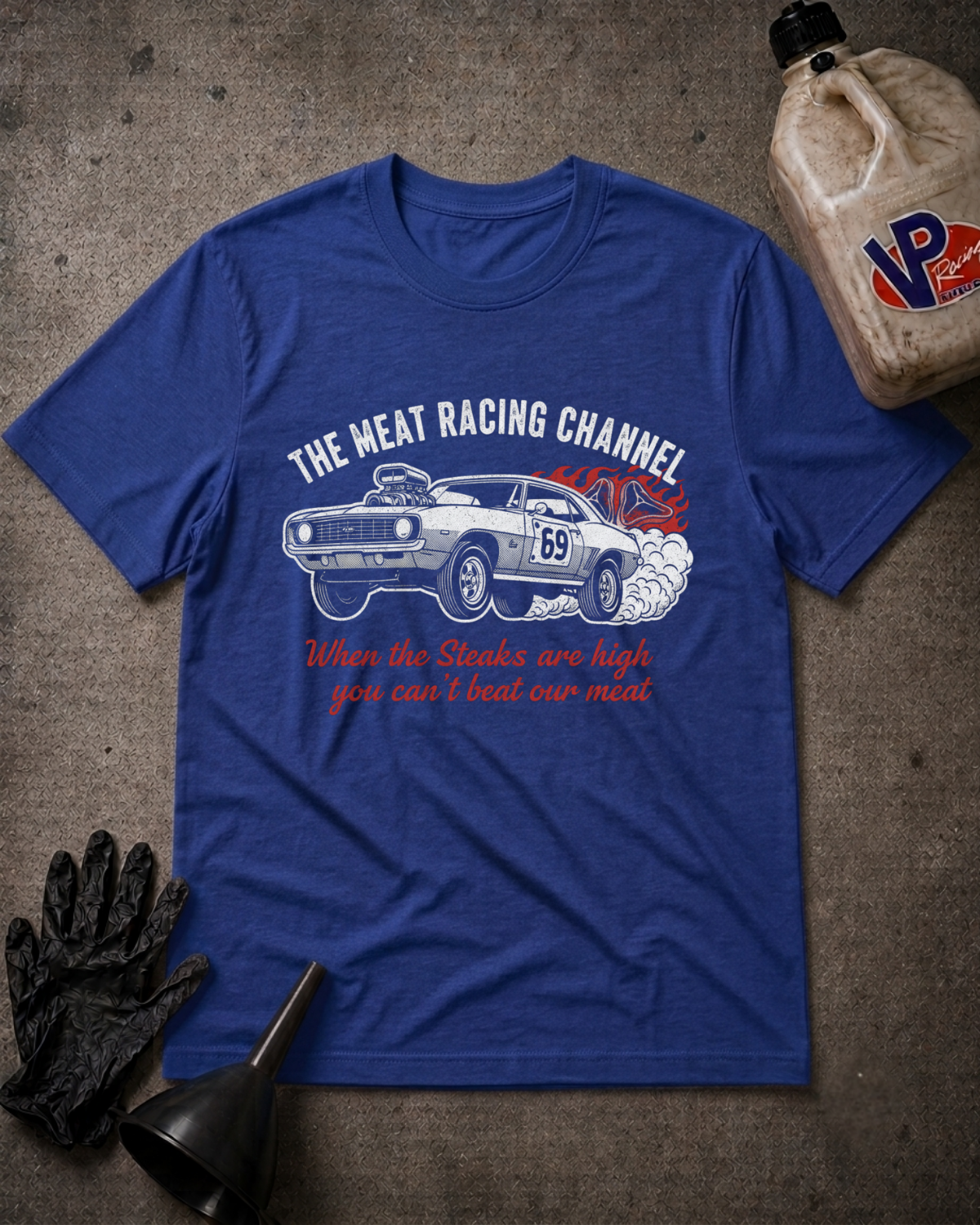 A royal blue graphic t-shirt laid flat on a textured gray surface, surrounded by racing-themed props including a bottle of VP Racing fluid, black gloves, and a funnel. The t-shirt features a white muscle car graphic with red smoke and the text 'THE MEAT RACING CHANNEL' and 'When the Steaks are high you can't beat our most'.