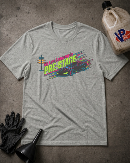 A heather gray graphic t-shirt laid flat on a textured gray surface, surrounded by automotive-themed props including a black glove, funnel, and a jug of oil. The shirt features a colorful graphic of a sports car and the text 'My Love Language is PRE-STAGE'.