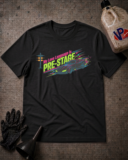 A black graphic t-shirt laid flat on a textured gray background, surrounded by automotive-themed props including a bottle of oil, a black glove, and a funnel. The shirt features a colorful graphic of a race car and the text 'My Love Language is PRE-STAGE'.