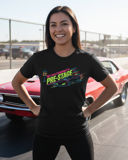 Woman wearing a black t-shirt with colorful text 'My Love Language is Pre-Stage' and graphic, standing in front of a red car on a track.