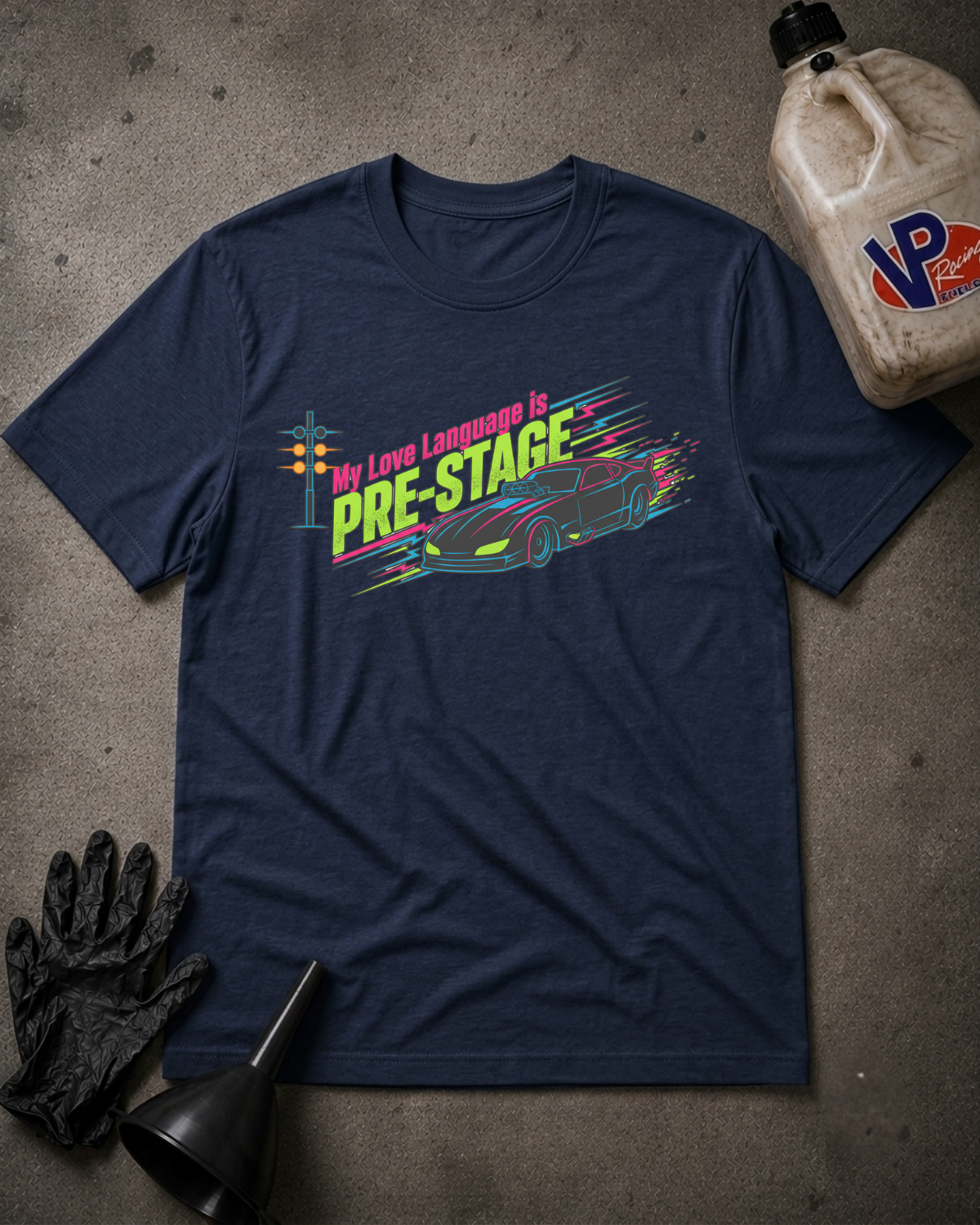 A navy blue graphic t-shirt laid flat on a gray concrete surface, surrounded by automotive-themed props including a black funnel, black gloves, and a plastic jug. The t-shirt features a colorful graphic of a race car and the text 'My Love Language is PRE-STAGE'.