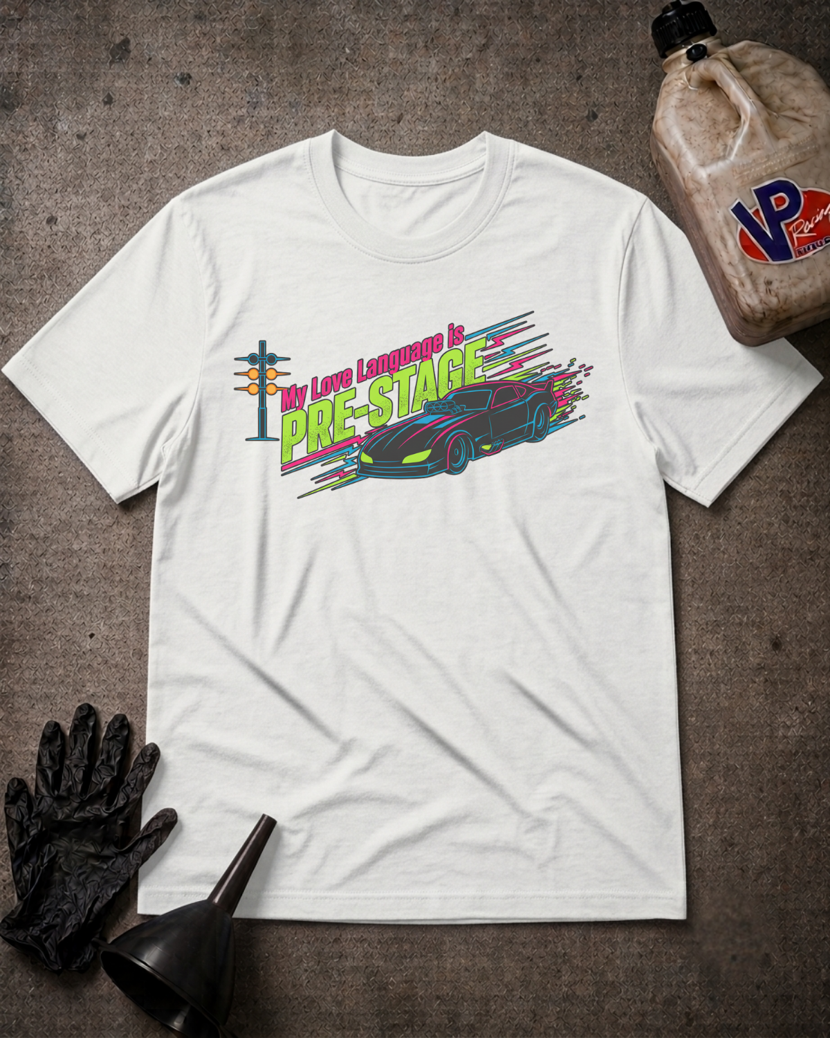 A white short-sleeve graphic T-shirt laid flat on a textured dark surface, surrounded by props including a black glove, black funnel, and a plastic jug. The shirt graphic reads 'The Love Language Is Pre-Stage' with a colorful car and neon-style lettering.
