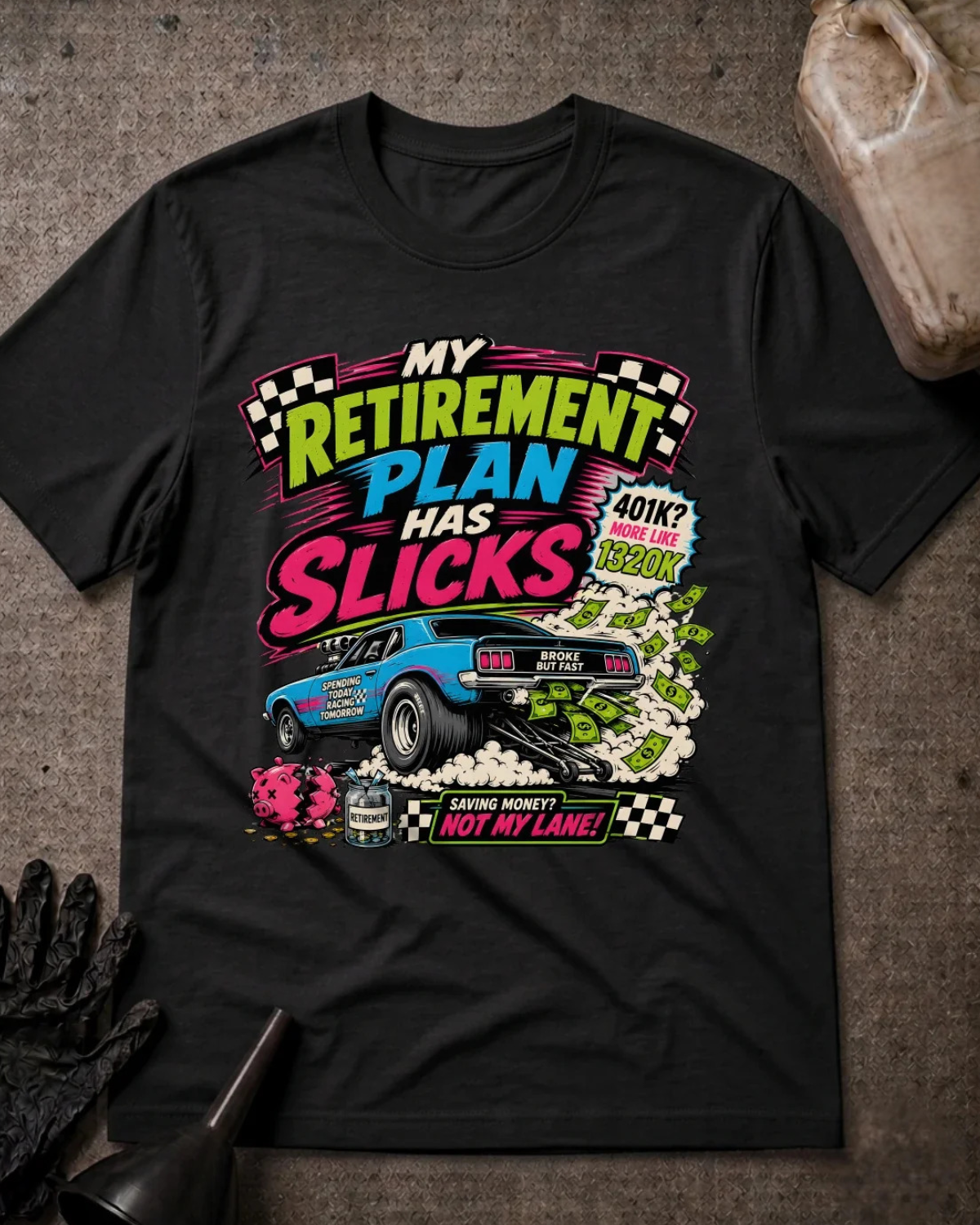 A black graphic t-shirt laid flat on a textured gray surface with racing-themed props like a glove, funnel, and bottle. The shirt features a colorful retro-style print with the text 'MY RETIREMENT PLAN HAS SLICKS' and a blue drag race car.