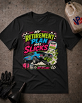A black graphic t-shirt laid flat on a textured gray surface, surrounded by racing-themed props including a black glove, funnel, and fuel canister. The shirt features a colorful graphic with the text 'MY RETIREMENT PLAN HAS SLICKS' and a blue race car.