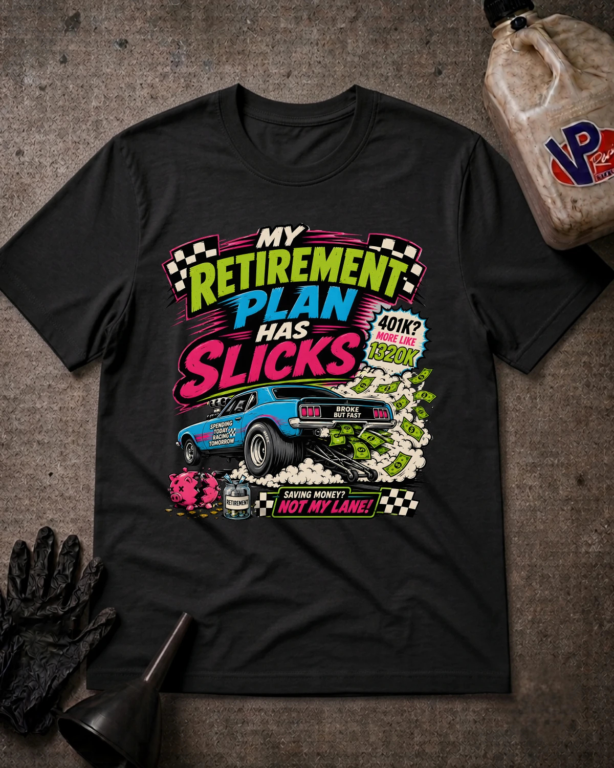 A black graphic t-shirt laid flat on a textured gray surface, surrounded by racing-themed props including a black glove, funnel, and fuel canister. The shirt features a colorful graphic with the text 'MY RETIREMENT PLAN HAS SLICKS' and a blue race car.