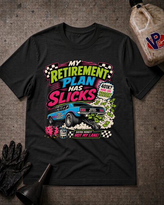A black graphic t-shirt laid flat on a textured gray surface, surrounded by racing-themed props including a black glove, funnel, and fuel canister. The shirt features a colorful graphic with the text 'MY RETIREMENT PLAN HAS SLICKS' and a blue race car.