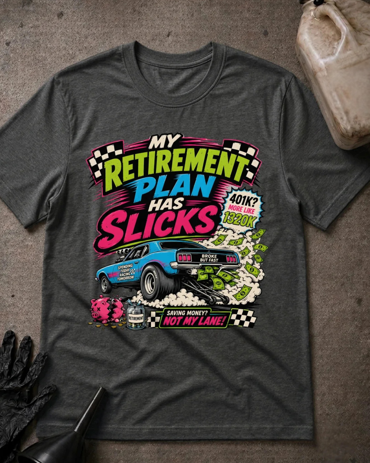 A dark heather gray graphic t-shirt laid flat on a textured gray surface, styled with a bottle cap, gloves, and a plastic jug. The shirt features a colorful graphic with the text 'MY RETIREMENT PLAN HAS SLICKS' and a blue muscle car surrounded by money and checkered flags.