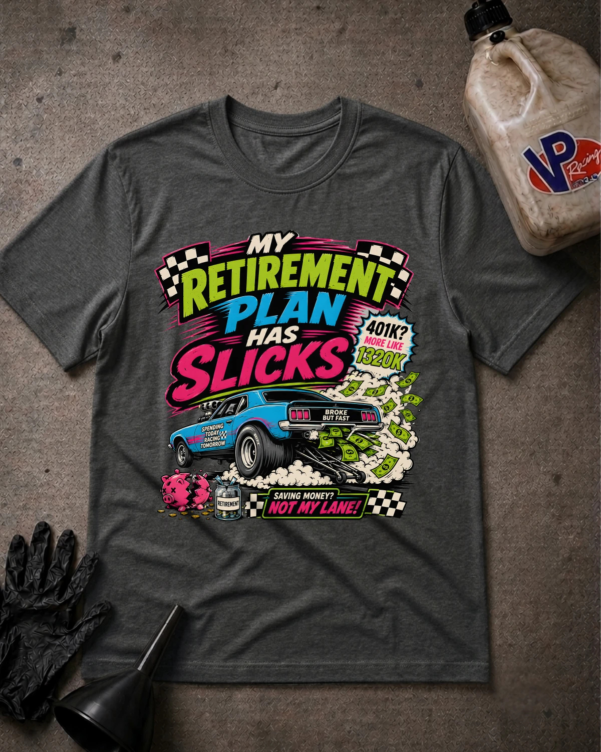 A dark heather gray graphic t-shirt laid flat on a textured gray surface, surrounded by racing-themed props including a black glove, funnel, bottle, and a beige jug with a VP logo. The t-shirt features a colorful retro-style graphic with the text 'MY RETIREMENT PLAN HAS SLICKS' and a blue muscle car.