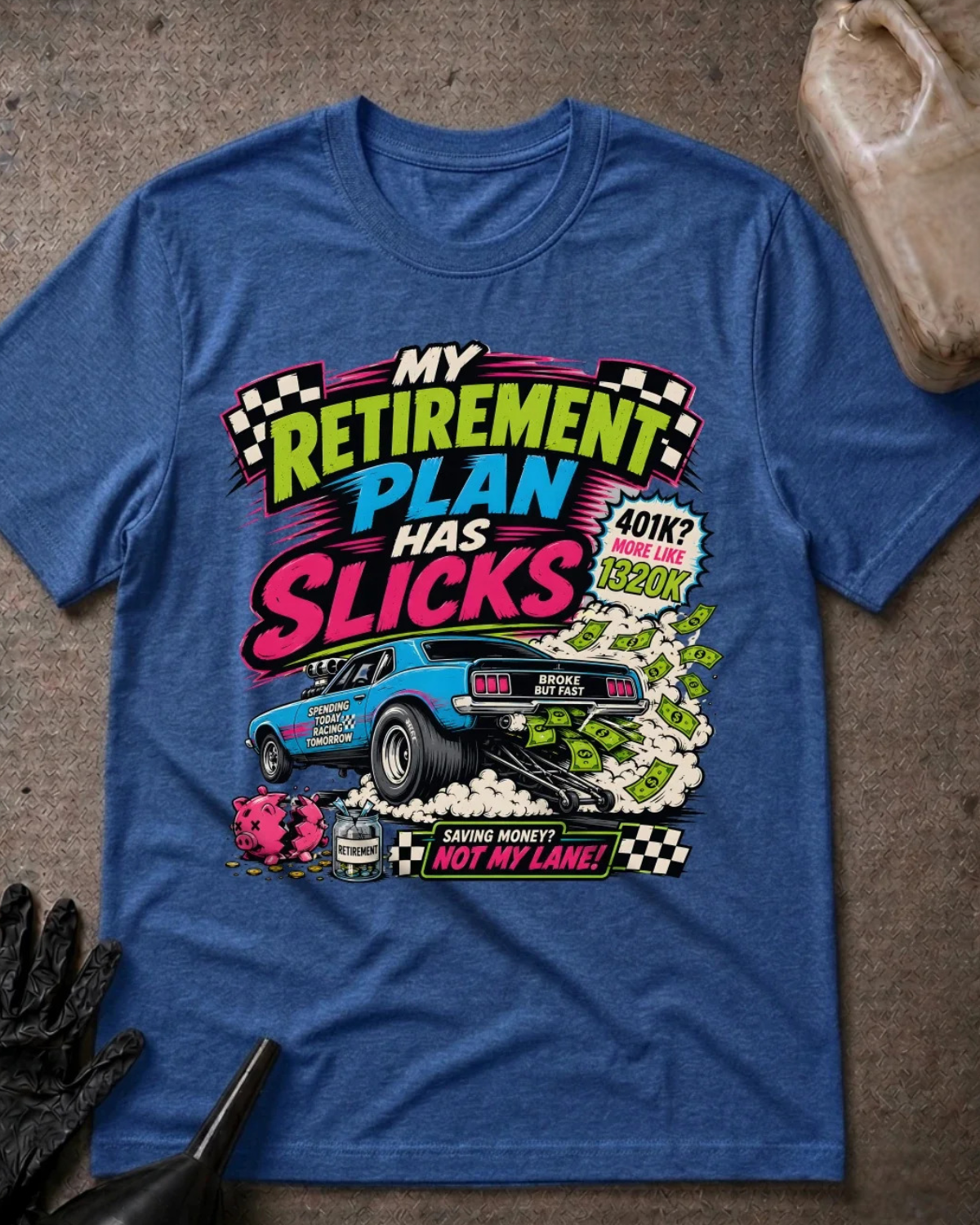 A heather blue graphic t-shirt laid flat on a textured gray surface, featuring a colorful racing-themed retirement slogan print that reads 'My Retirement Plan Has Slicks' with a blue race car, dollar bills, and checkered flags.