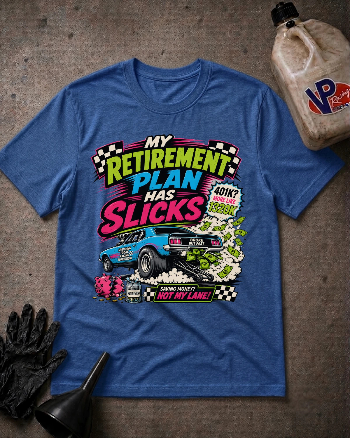 A blue graphic t-shirt laid flat on a textured gray surface, surrounded by automotive-themed props including a black glove, a funnel, and a bottle of oil. The t-shirt features a colorful retro-style comic graphic with the text 'MY RETIREMENT PLAN HAS SLICKS' and a blue race car.