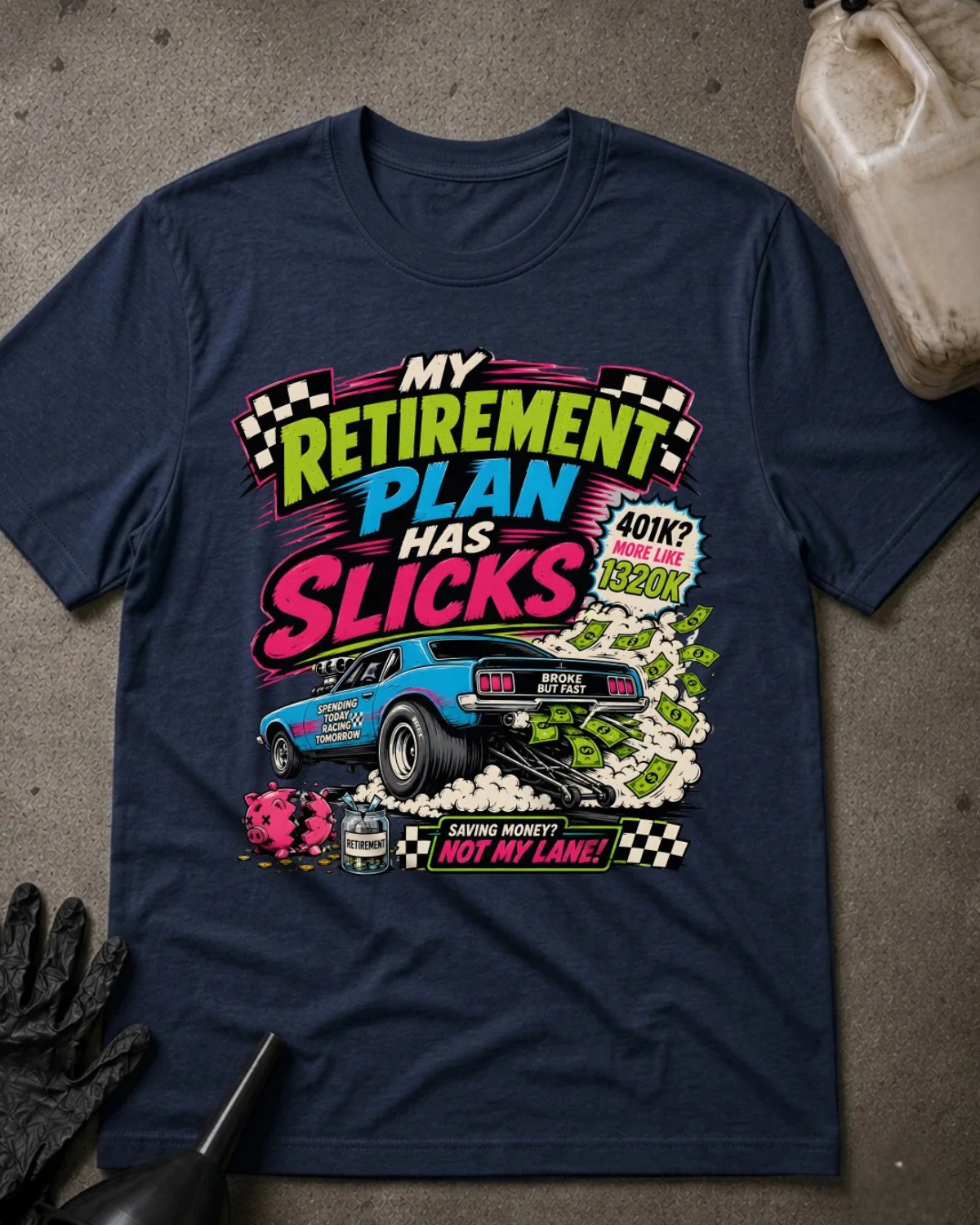 A navy blue graphic t-shirt laid flat on a gray concrete-like surface, featuring a colorful race car and bold text that reads 'My Retirement Plan Has Slicks' with additional racing-themed graphics. A black glove, a drill bit, and part of a plastic jug are visible around it as styled props.