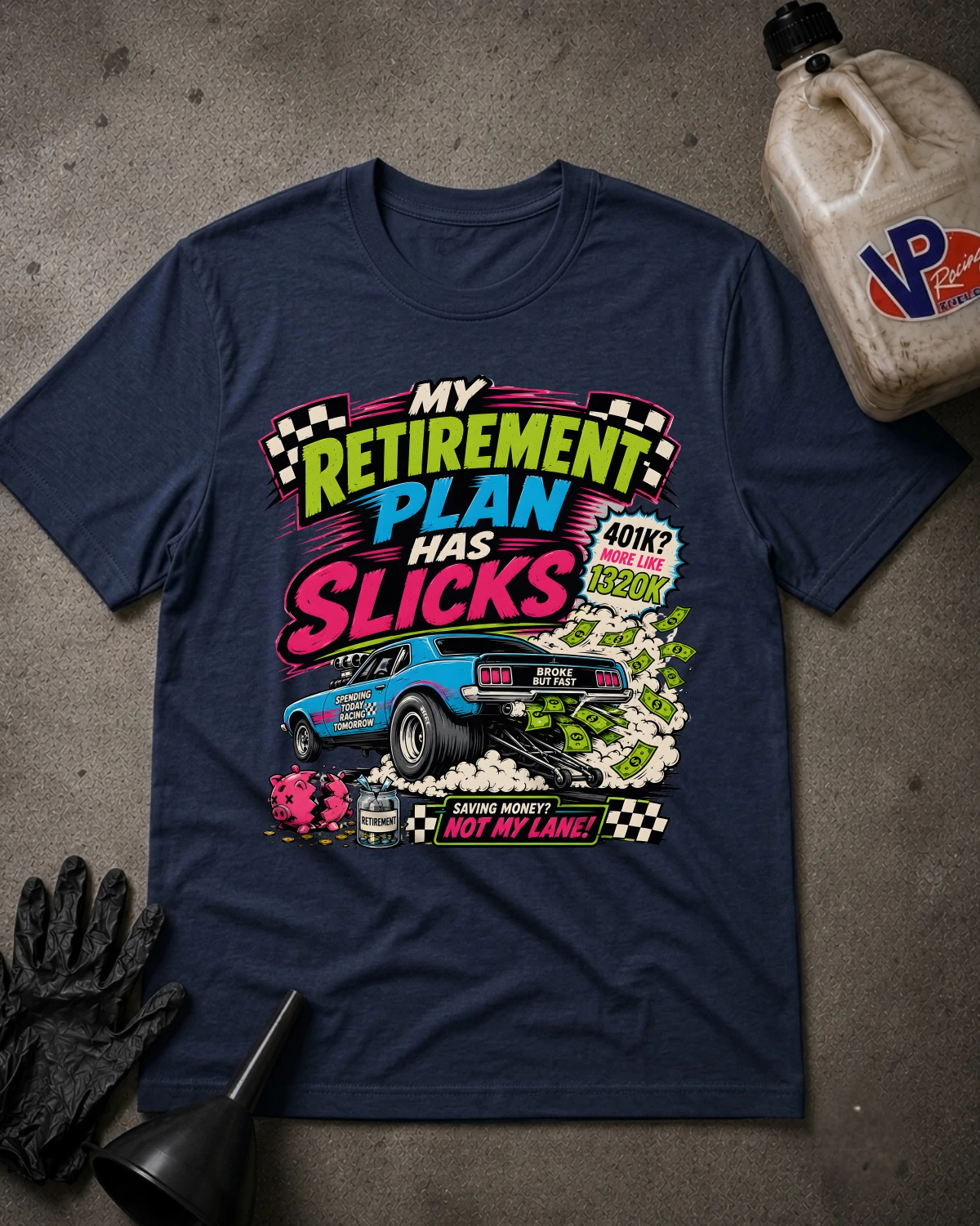 A navy blue graphic t-shirt laid flat on a gray concrete-like surface, styled with black gloves, a funnel, and a plastic jug. The shirt features a colorful retro-style car wash advertisement graphic with the text 'My Retirement Plan Has Slicks' and 'Saving Money Not My Land!'.