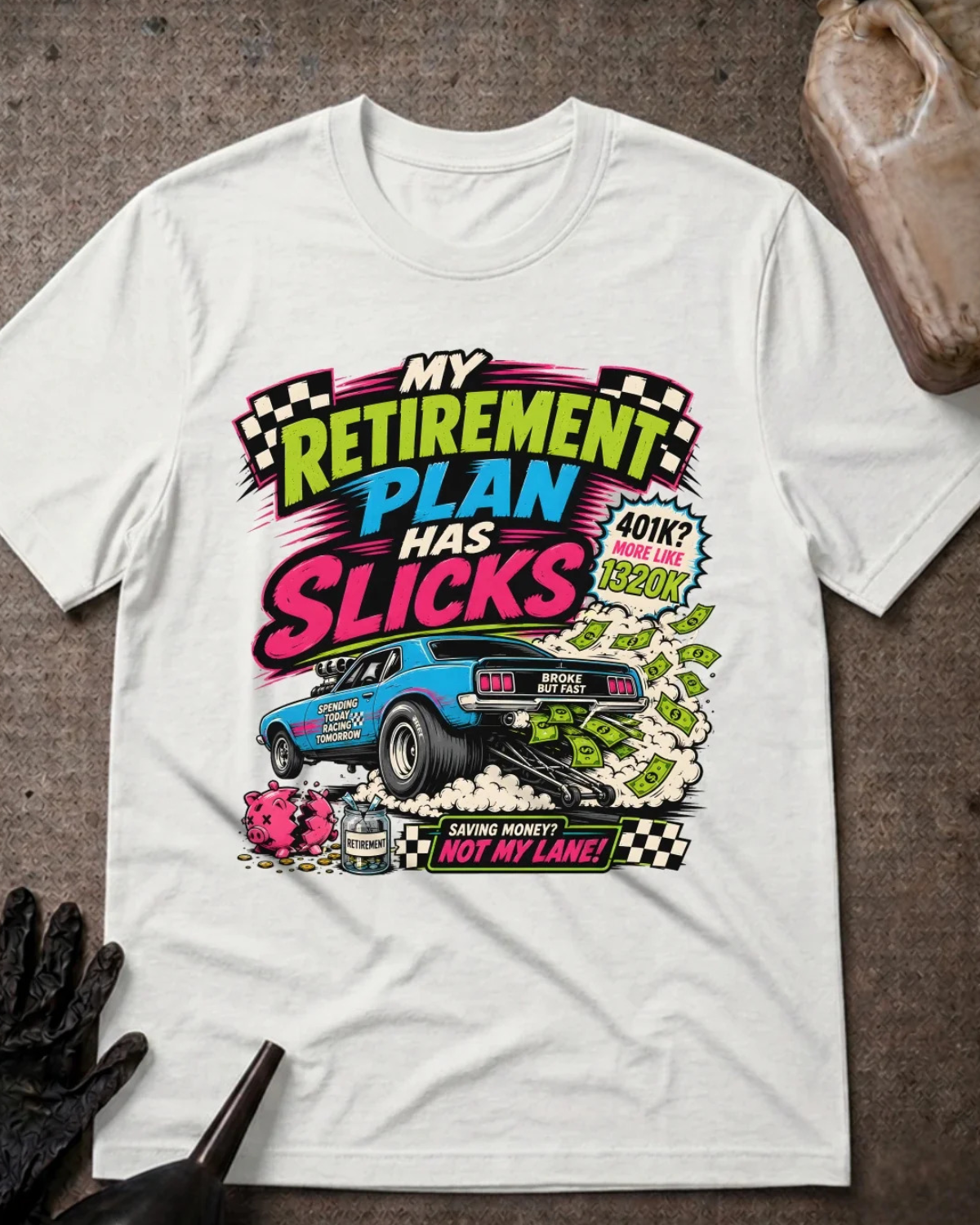 A white short-sleeve graphic T-shirt laid flat on a textured dark surface. The shirt features a colorful retro-style racing graphic with the text 'My Retirement Plan Has Slicks' and a blue race car, plus small props including a black glove and a stone in the corners.