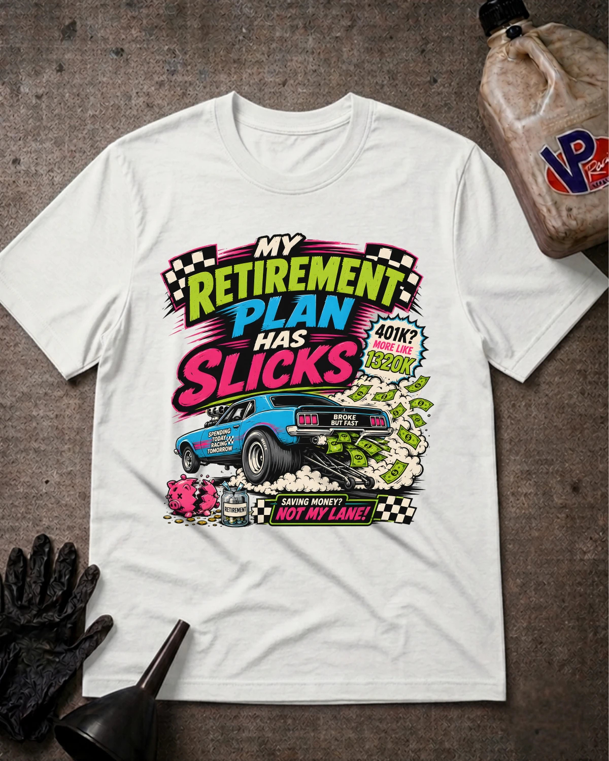 A white short-sleeve graphic T-shirt laid flat on a textured gray surface, surrounded by racing-themed props including a jug, gloves, and a funnel. The shirt features a large colorful retro-style print reading 'My Retirement Plan Has Slicks' with a blue race car, money bags, and checkered details.