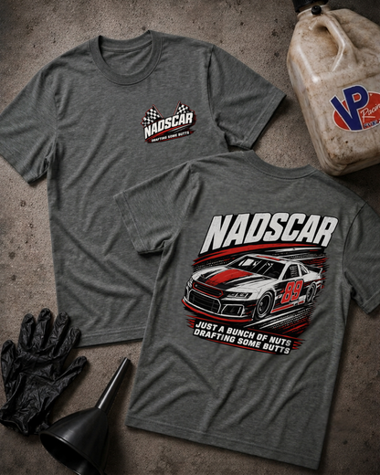 Two heather gray graphic T-shirts laid flat on a textured concrete surface, with a NADSCAR chest logo on the front shirt and a large NADSCAR race car graphic on the back shirt. Black gloves and a funnel sit near the shirts, and a dirty plastic jug with a VP logo is at the top right.