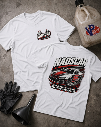 Two white graphic t-shirts displayed flat on a gray concrete surface, with a NADSCAR logo on the front and a large NADSCAR race car graphic on the back. Styled flat-lay props include a plastic jug, black gloves, and a funnel.