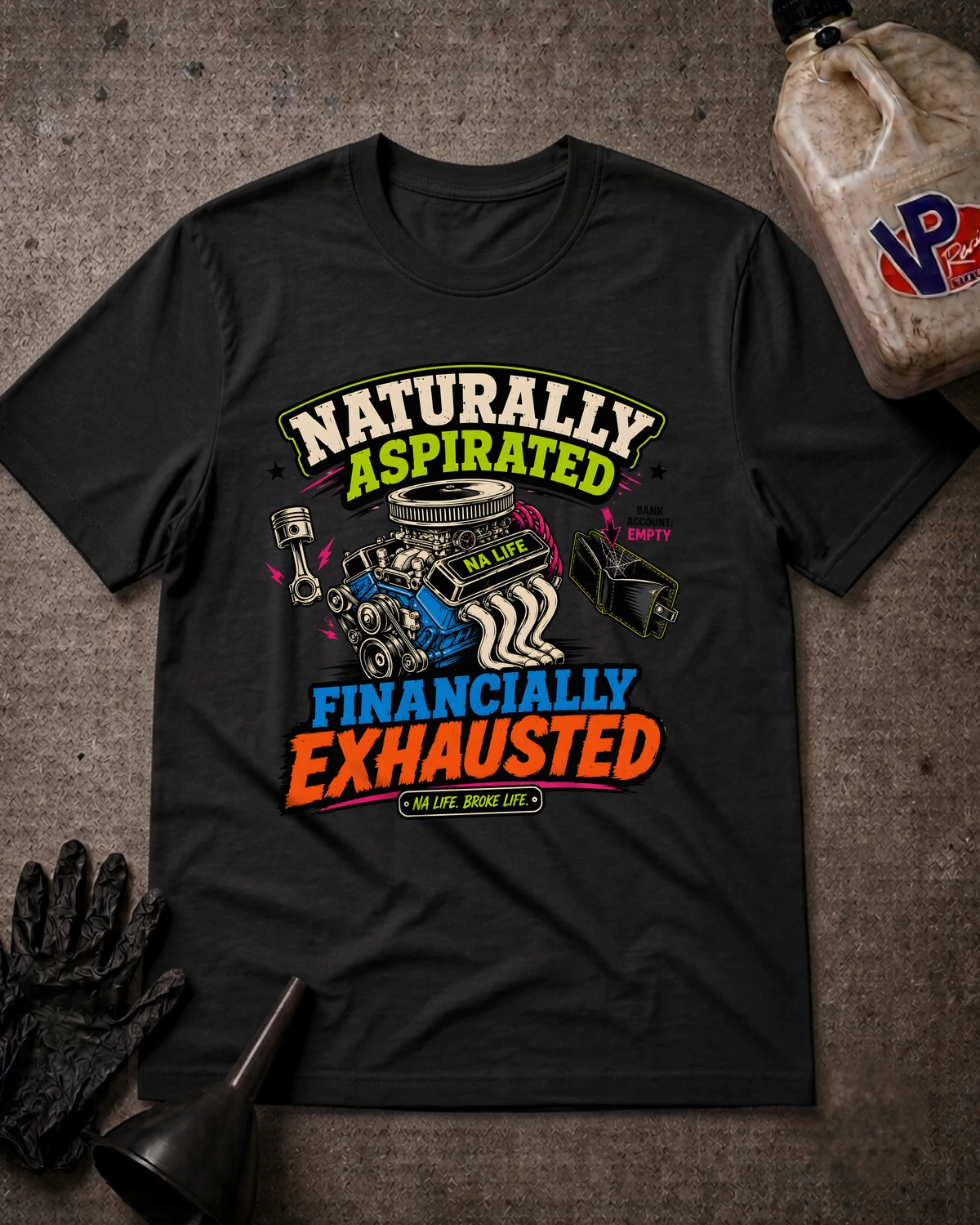 A black graphic t-shirt laid flat on a textured gray surface, surrounded by automotive-themed props including a plastic jug, gloves, and a funnel. The shirt features a colorful graphic with the text 'NATURALLY ASPIRATED FINANCIALLY EXHAUSTED' and illustrations of an engine, pistons, and a steering wheel.