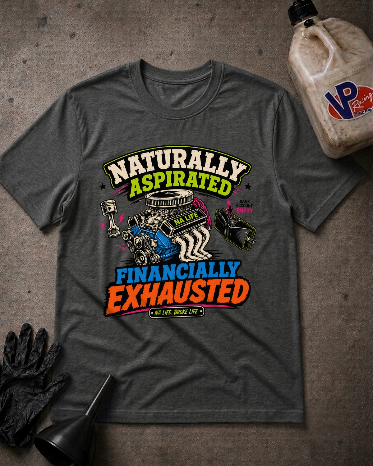 A dark heather gray graphic t-shirt laid flat on a textured gray surface, surrounded by automotive-themed props including a bottle of oil, a black glove, and a funnel. The t-shirt features a colorful graphic with the text 'Naturally Aspired Financially Exhausted' and illustrations of a car engine, pistons, and a wrench.