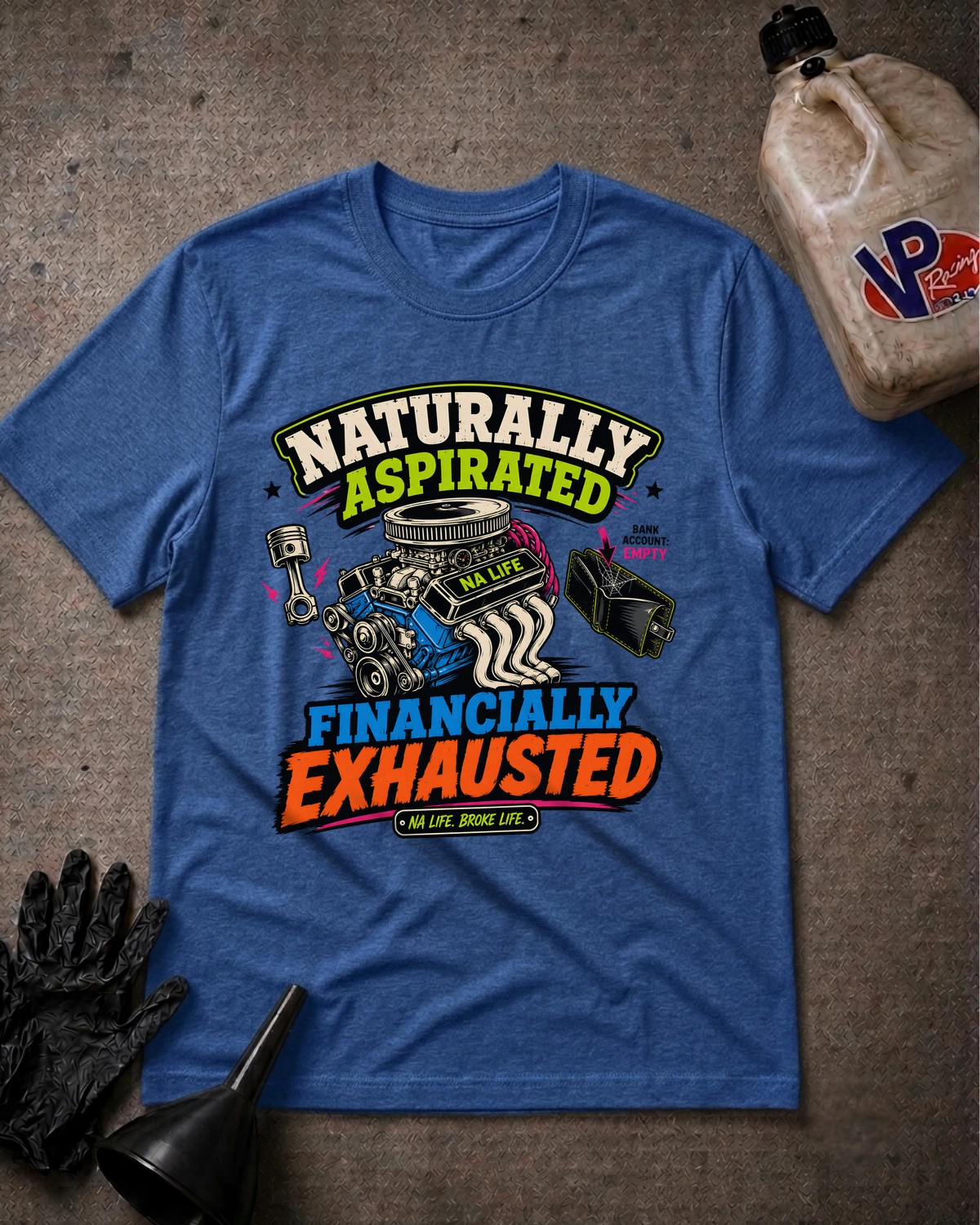 A heather blue graphic t-shirt laid flat on a textured gray surface, surrounded by automotive-themed props including a bottle of oil, a black glove, and a funnel. The shirt features a colorful graphic with an engine illustration and the text 'Naturally Aspirated FINANCALLY EXHAUSTED'.
