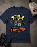 A navy blue graphic t-shirt laid flat on a gray concrete-like surface, surrounded by black gloves, a funnel, and a plastic jug. The shirt features a colorful graphic with an engine illustration and the text 'NATURALLY ASPIRATED FINANCALLY EXHAUSTED' with 'NO LIFE ENGINE LIFE' underneath.