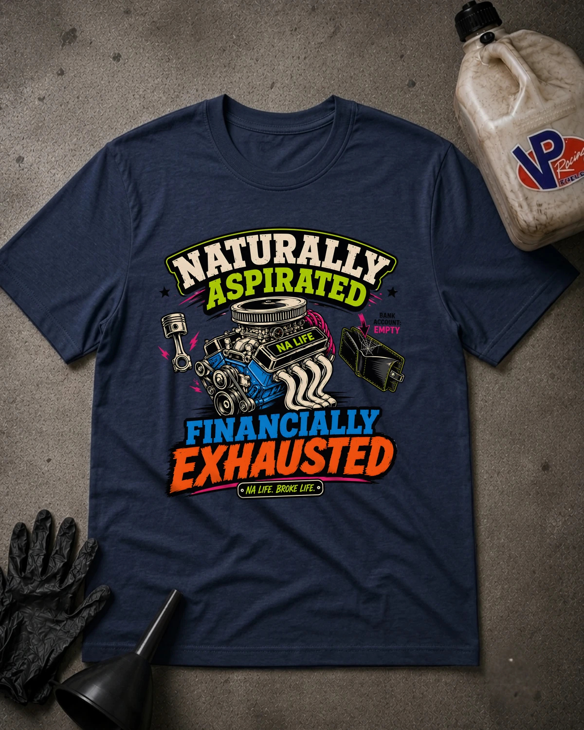 A navy blue graphic t-shirt laid flat on a gray concrete-like surface, surrounded by black gloves, a funnel, and a plastic jug. The shirt features a colorful graphic with an engine illustration and the text 'NATURALLY ASPIRATED FINANCALLY EXHAUSTED' with 'NO LIFE ENGINE LIFE' underneath.