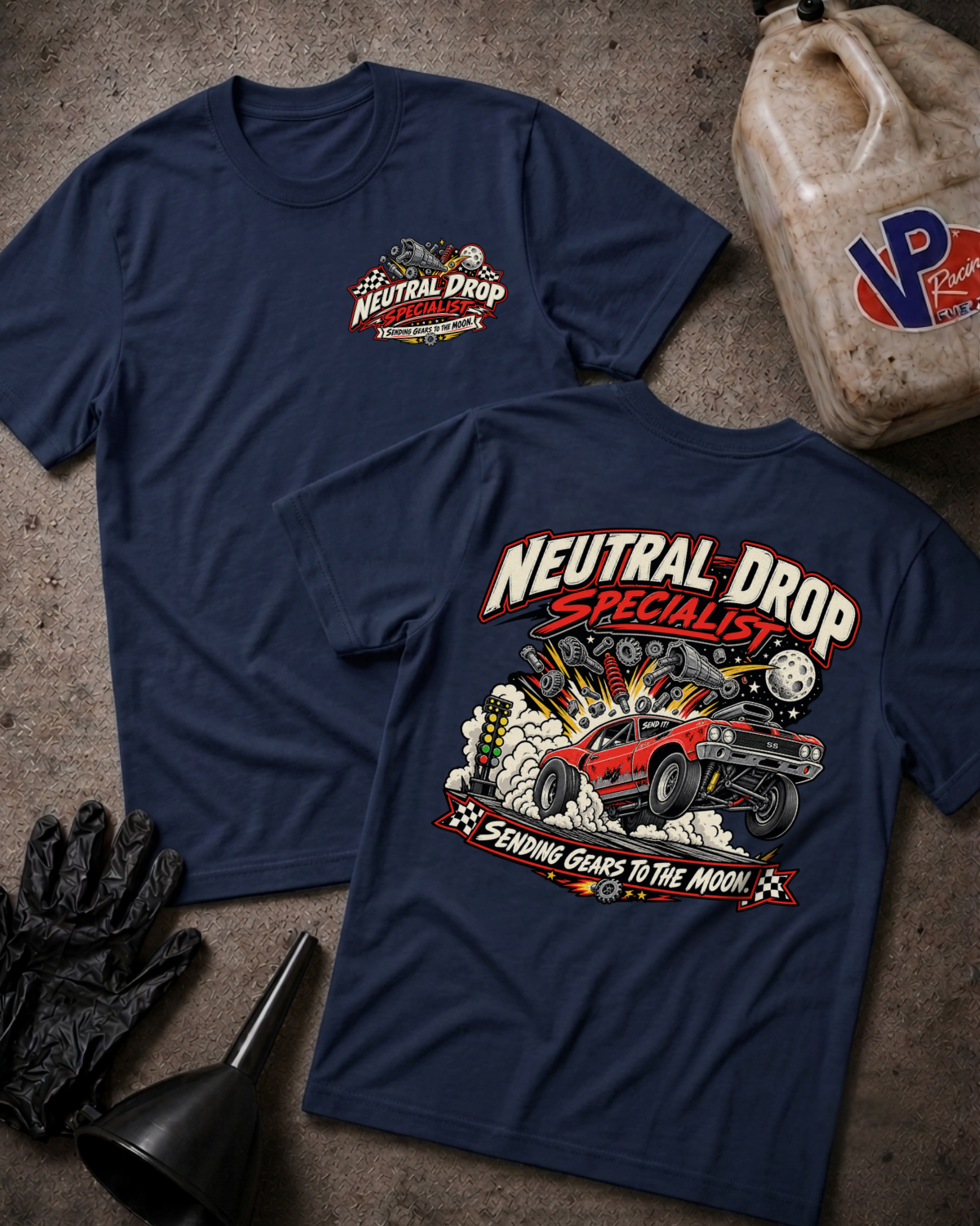 Two navy blue graphic T-shirts laid flat on a textured gray surface, with automotive-themed printed artwork on the chest and back with 'NEUTRAL DROP SPECIALIST SENDING GEARS TO THE MOON' text. A plastic jug, black gloves, and a funnel are placed around them as styled props.