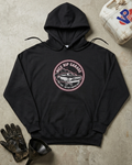 A black pullover hoodie laid flat on a textured concrete surface, featuring a circular vintage-style garage graphic on the chest with the words 'Nice Rip Garage' and a car illustration. Surrounding props include a worn work glove, funnel, and a dirty fuel can.