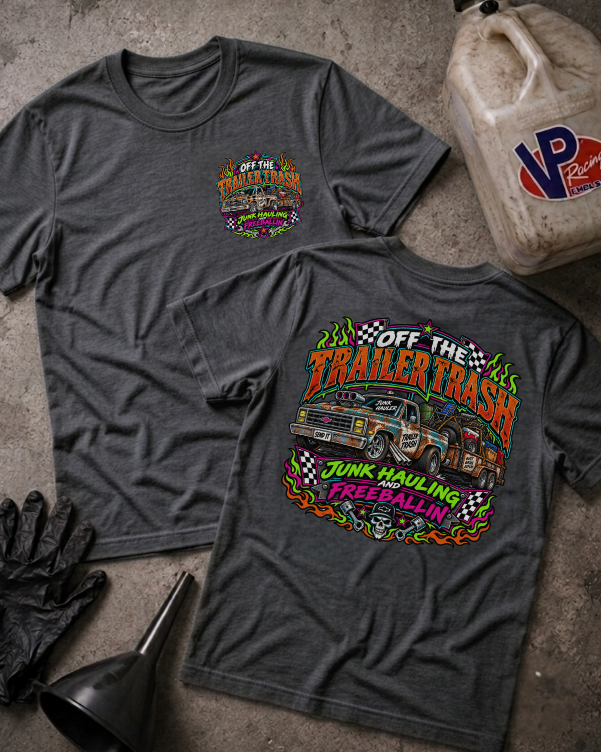 Two dark heather gray graphic t-shirts displayed flat on a concrete surface, with a black funnel, black gloves, and a VP gas can nearby. The front shirt shows a small colorful chest graphic, and the back shirt shows a large colorful graphic reading 'Off The Trailer Trash Junk Hauling and Freeball' with a truck illustration.
