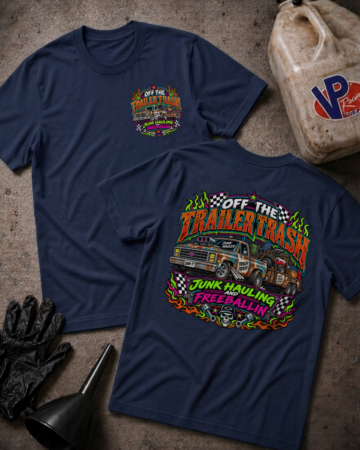 Two navy blue graphic t-shirts displayed on a textured concrete surface with racing-themed props including a fuel can, gloves, and funnel. The front shirt shows a small chest graphic, while the back shirt features a large colorful graphic of a race truck and the text 'Off The Trailer Trash Junk Hauling and Freeballin'