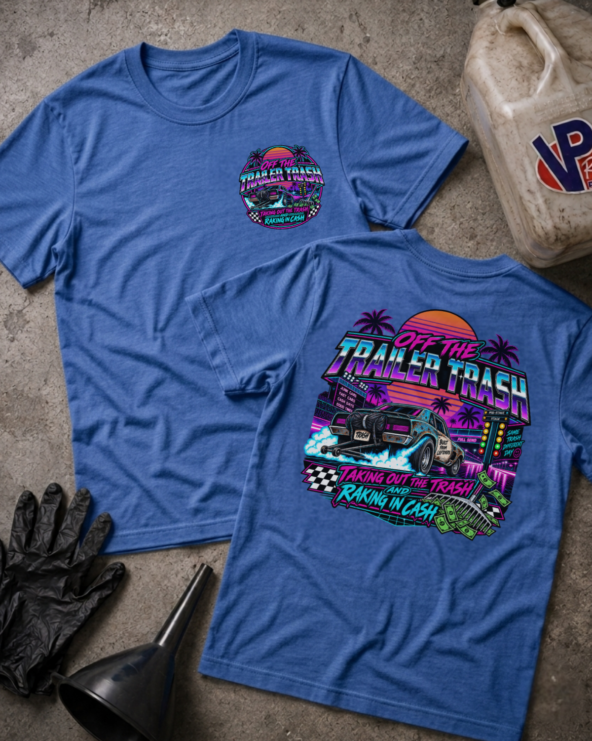Two heather true royal graphic t-shirts displayed flat on a textured concrete surface, surrounded by garage-themed props including a jerrycan, black gloves, and a funnel. The front shirt features a small circular logo on the left chest, while the back shirt displays a large, colorful retro-style graphic with a monster truck and the text 'OFF THE TRAILER TRASH TAKING OUT THE TRASH RAKIN' IN CASH'.