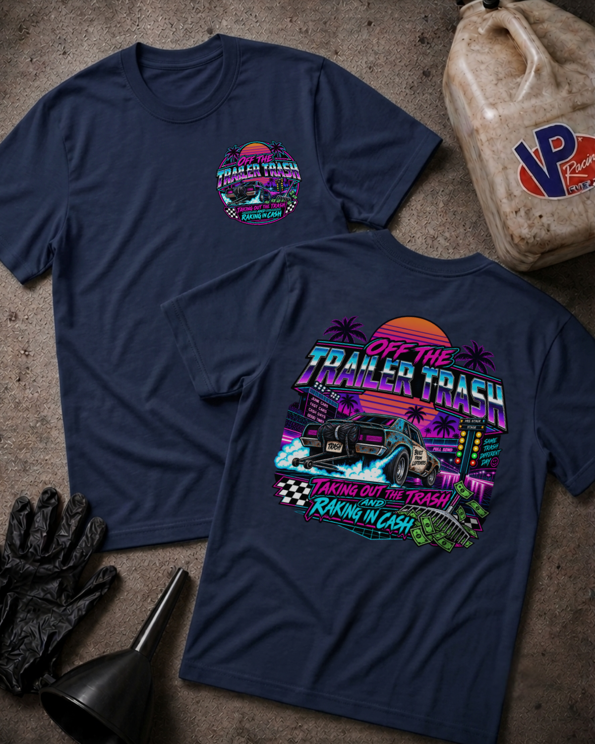 Two navy blue graphic t-shirts displayed flat on a textured concrete surface, surrounded by garage-themed props including a jerrycan, black gloves, and a funnel. The front shirt features a small circular logo on the left chest, while the back shirt displays a large, colorful retro-style graphic with a monster truck and the text 'OFF THE TRAILER TRASH TAKING OUT THE TRASH RAKIN' IN CASH'.