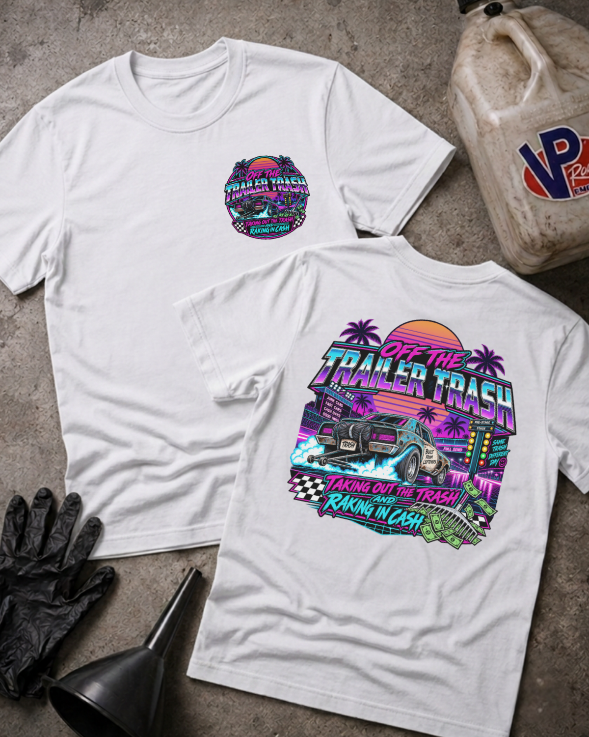 Two white graphic t-shirts displayed flat on a textured concrete surface, surrounded by garage-themed props including a jerrycan, black gloves, and a funnel. The front shirt features a small circular logo on the left chest, while the back shirt displays a large, colorful retro-style graphic with a monster truck and the text 'OFF THE TRAILER TRASH TAKING OUT THE TRASH RAKIN' IN CASH'.